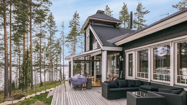 Um deck de madeira com mobiliário externo, com vista para um lago cercado por árvores altas.