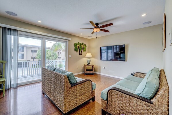 A spacious living room with wicker furniture, a ceiling fan, and a view through a sliding glass door to the outside.