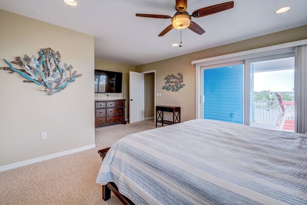 A bedroom with a bed, a ceiling fan, a dresser, a TV, and a door leading to a balcony.