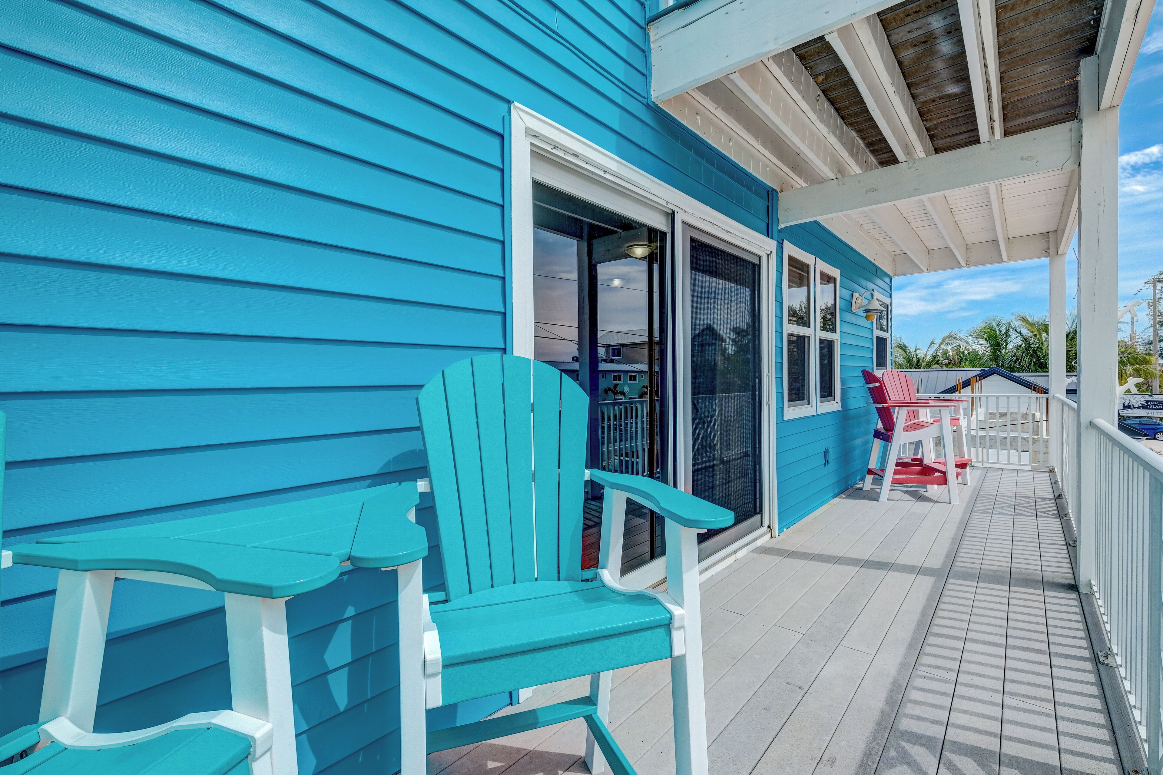 Une véranda couverte avec des fauteuils Adirondack turquoise, une table blanche et un bâtiment bleu avec une porte blanche.