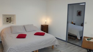 A bedroom with a bed, a nightstand, a red pillow, a small table, and a door leading to another room. - Gîte 4 persons; Autour d'un Puits (Marsilly)