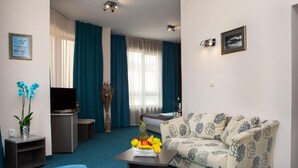 Habitación de hotel con cama, sofá, mesa de centro con fruta, televisión y ventana con cortinas.
