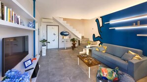 Una sala de estar moderna con un televisor de pantalla plana, una estantería, un sofá azul, una mesa de centro y una escalera que conduce a otro nivel.