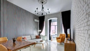 A modern living room with a wooden dining table, sofa, and armchairs, a white brick wall, and a large window with curtains.