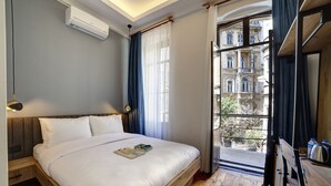 A bedroom with a wooden bed, a nightstand, a reading lamp, a window with curtains, and a view of a building outside.