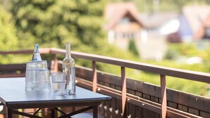Uma mesa com copos e garrafas de vidro transparente em um deck externo com guarda-corpo de madeira, com vista para uma paisagem verde e casas.