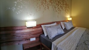 A hotel room with a wooden headboard, two bedside lamps, a bed with patterned pillows, and a wall with floral wallpaper. - 45C Coco Canopy Sea views Hiking Kiting Beaches Adventure (La Gaulette)