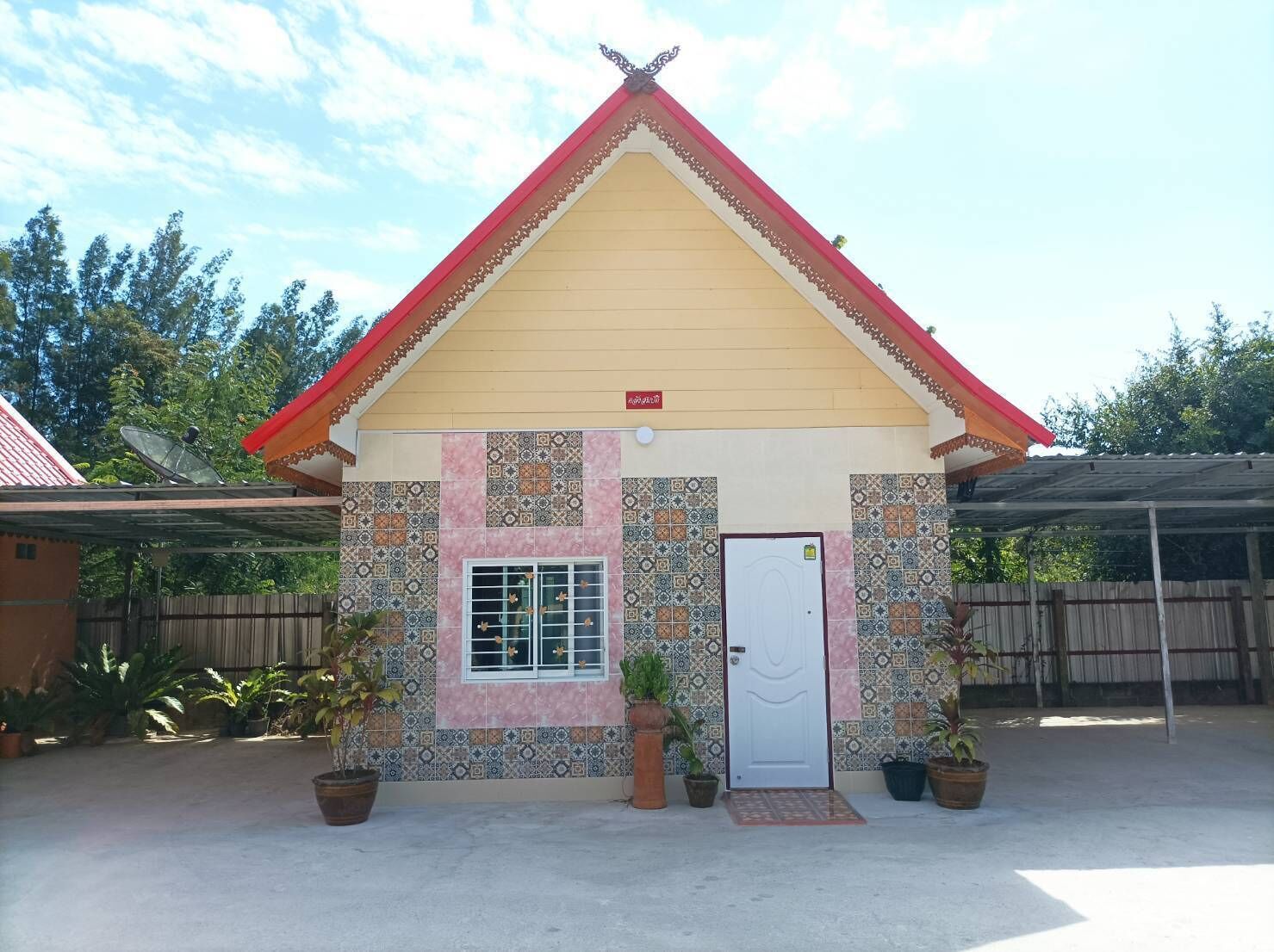 A small building with a red roof and a white door, surrounded by potted plants and a wooden fence.