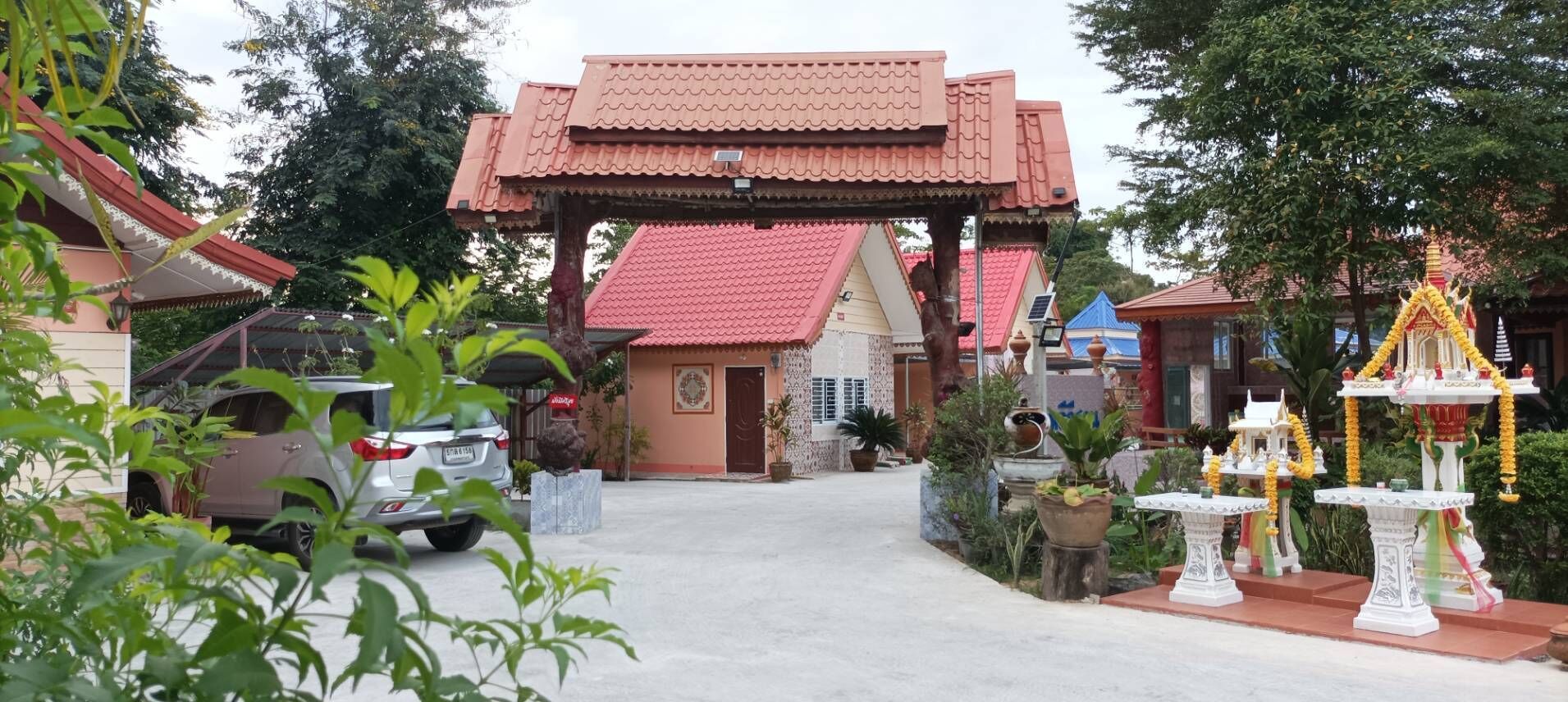 Ein traditioneller thailändischer Tempel mit einem pavilIon mit rotem Dach, einem parkenden Auto und einem kleinen Schrein mit Gaben.