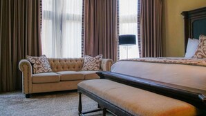 A hotel room with a large bed, a sofa, and a bench. There are curtains on the windows, and a lamp is visible.