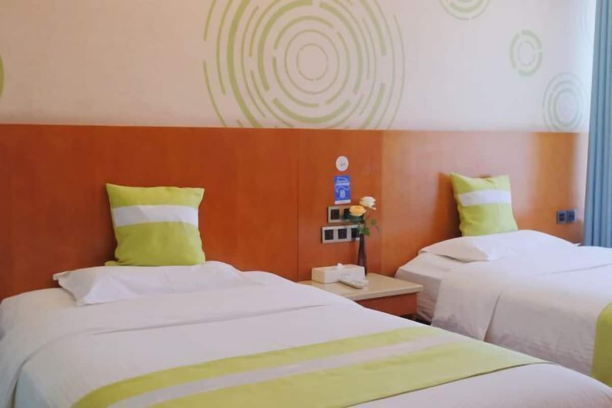 A hotel room with two beds, each with white bedding and green accents. There is a wooden headboard, a nightstand with a phone and a vase with a flower, and a wall with circular patterns.