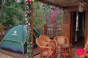 A wooden deck with a green tent labeled 'CAMELSPORTS,' wicker chairs, a small table, and a hanging sculpture.