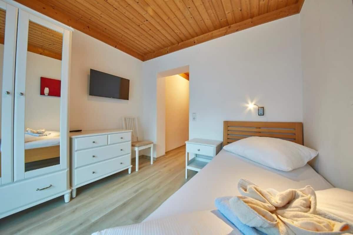 A bedroom with a wooden ceiling, a bed with white linens, a white wardrobe, a bedside table, and a small chair.