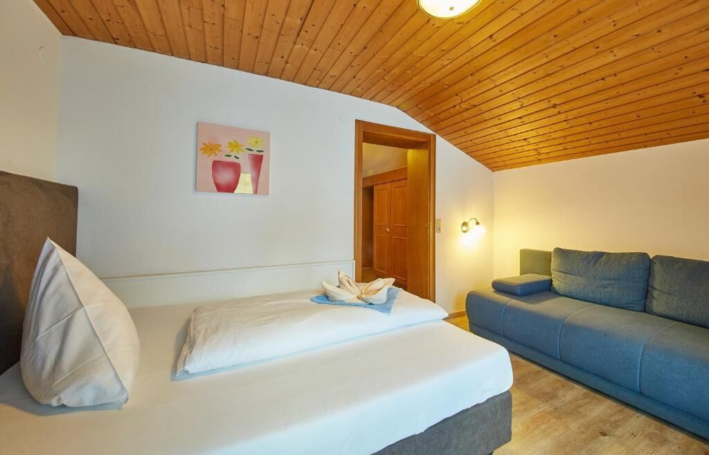 A bedroom with a bed, a blue sofa, a wooden ceiling, and a door leading to another room.