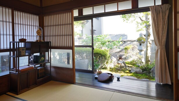 A traditional Japanese room with tatami flooring, a wooden shelf with various items, a large window with a view of a garden, and a sliding door. - Uji Tea Inn (Uji)