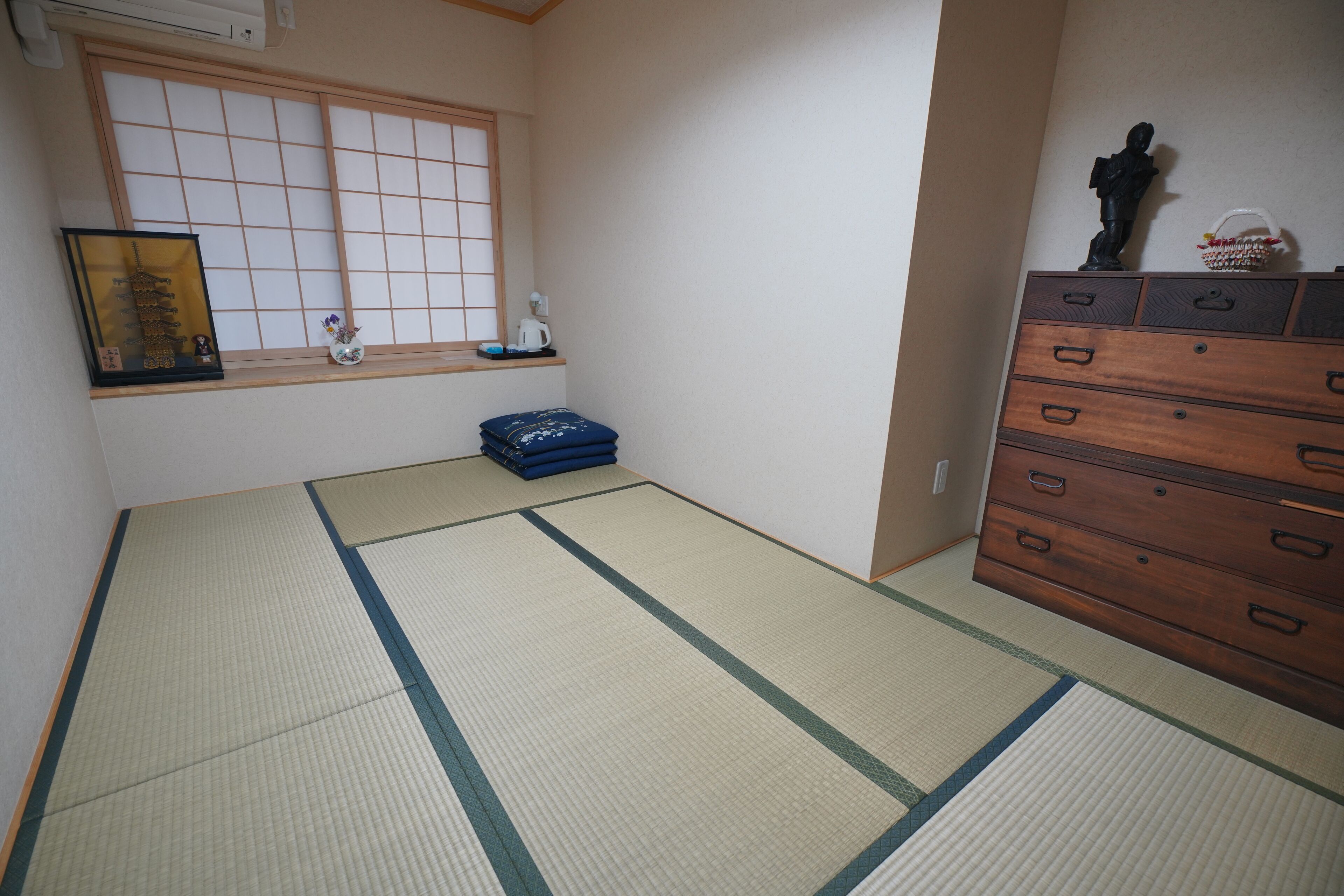 Une pièce avec des tatamis, une commode en bois, une fenêtre ornée de paravents shoji et une petite étagère sur laquelle sont disposés des objets décoratifs.
