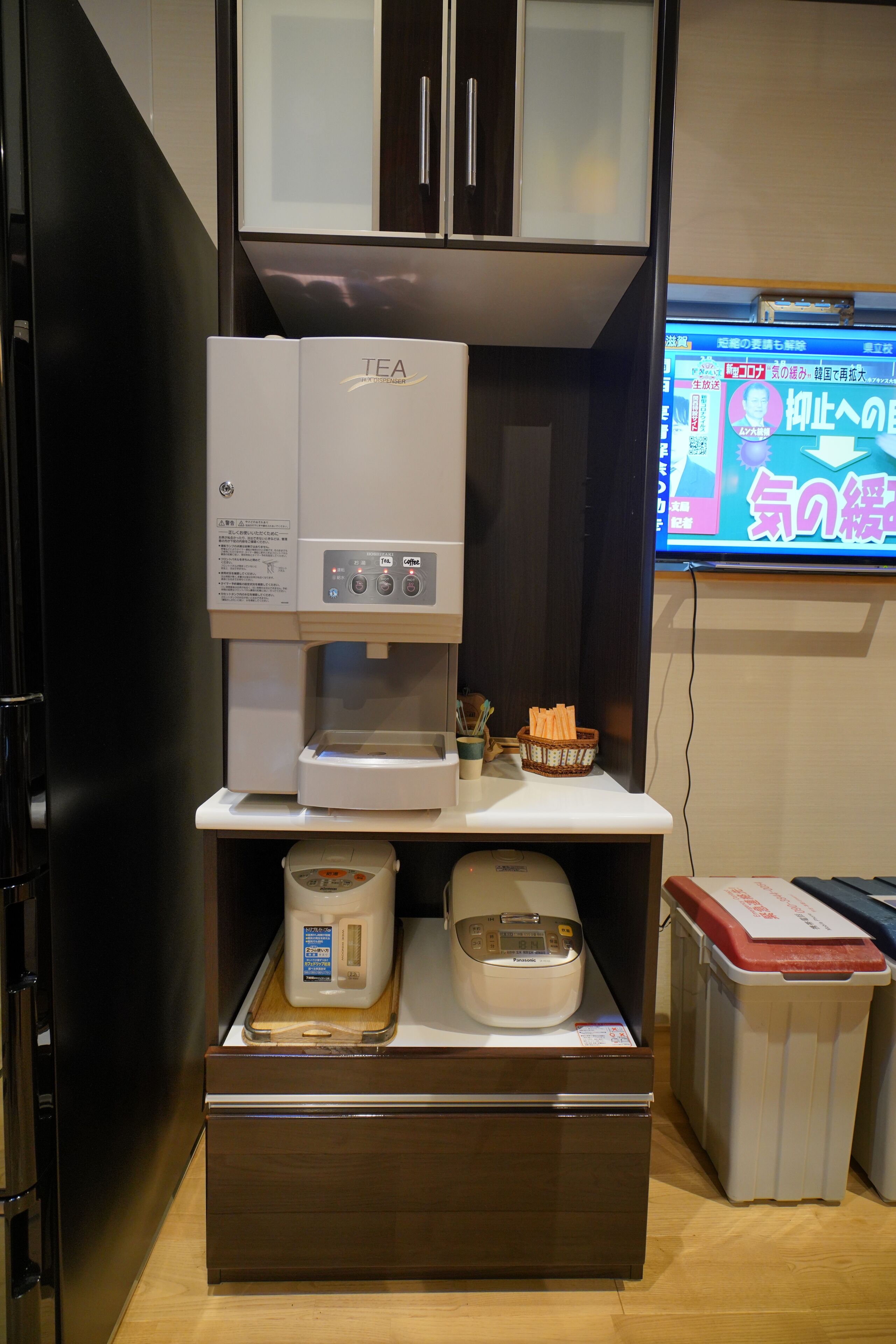 A kitchen area with a coffee machine, a microwave, and a refrigerator. There is a television mounted on the wall displaying Japanese text.