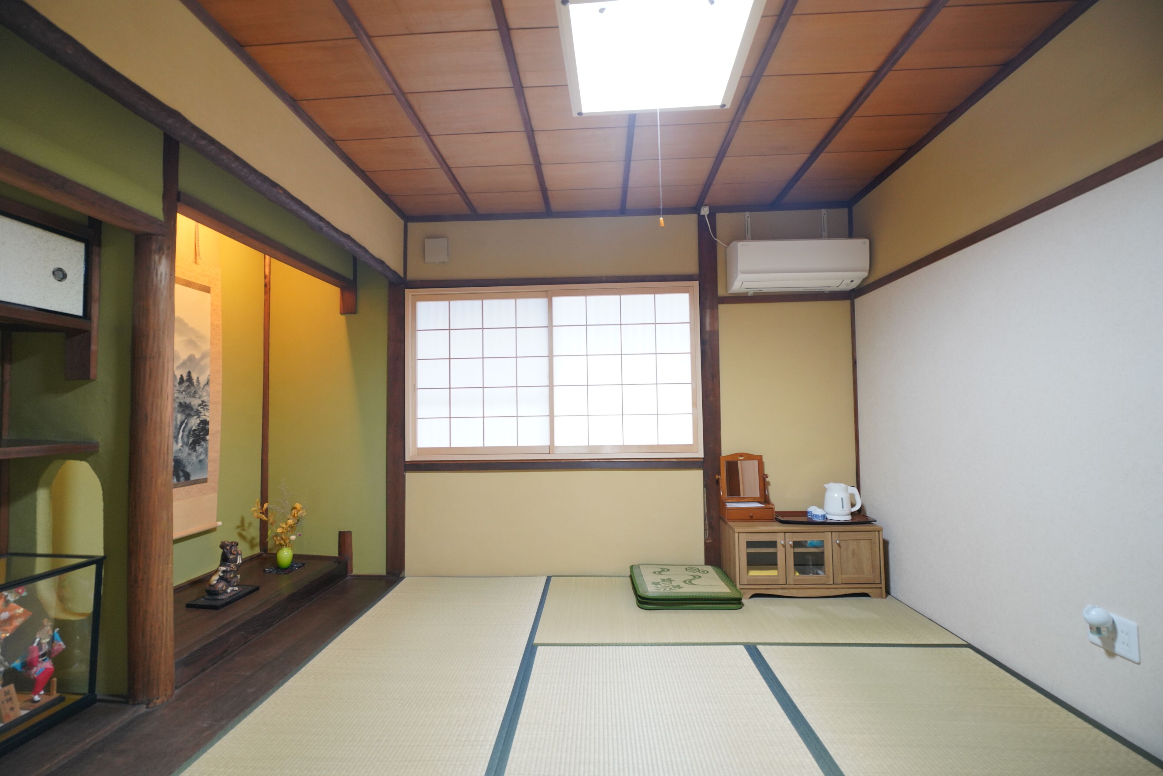 Une pièce avec un sol recouvert de tatamis, une porte coulissante, une fenêtre, un petit meuble avec une bouilloire et une étagère où sont disposés des objets décoratifs.