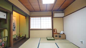 Una habitación con tatamis, una puerta corredera, una ventana, un mueble pequeño con hervidor y un estante con adornos.