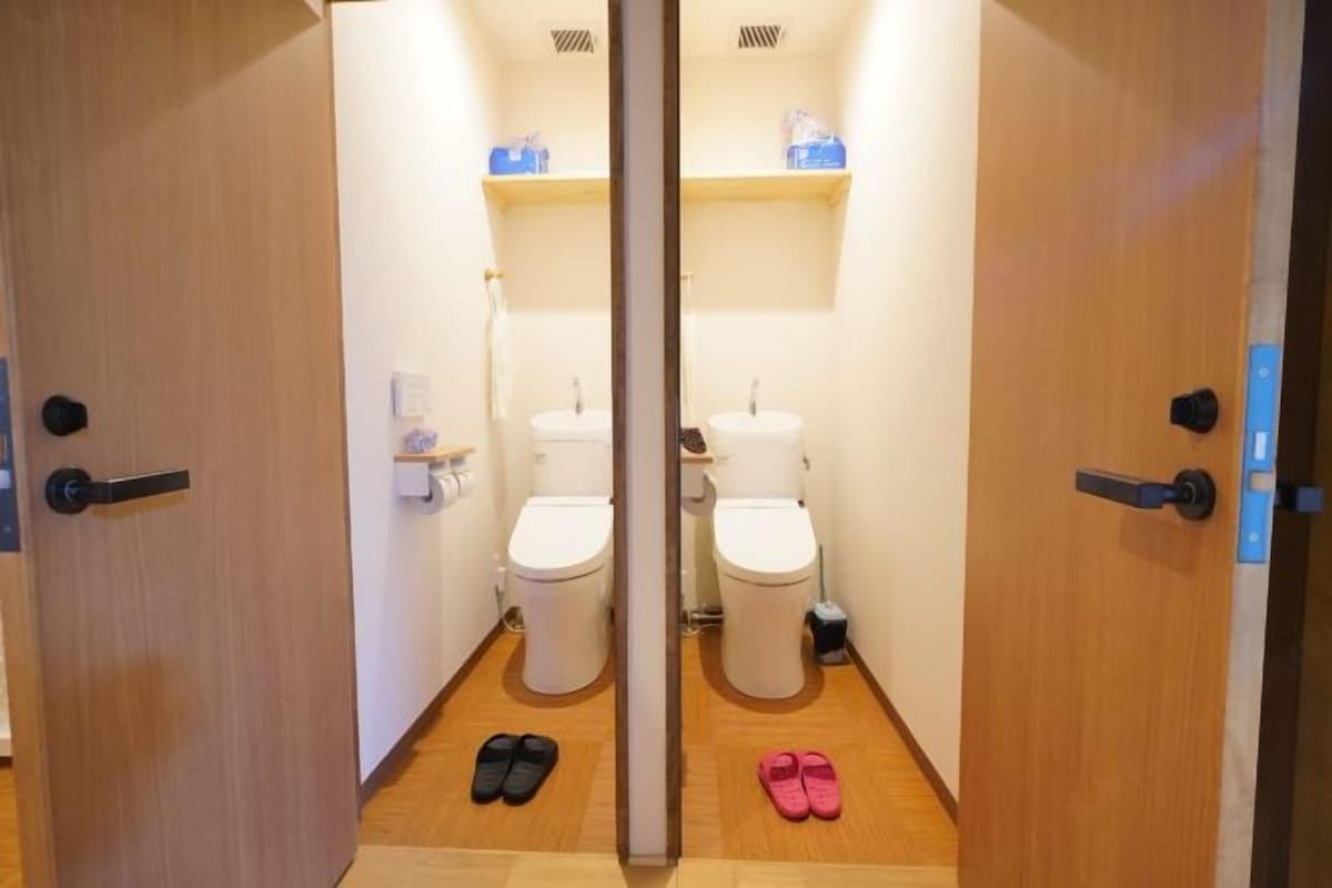 A bathroom with two toilets, a sink, and a mirror. There are slippers on the floor.