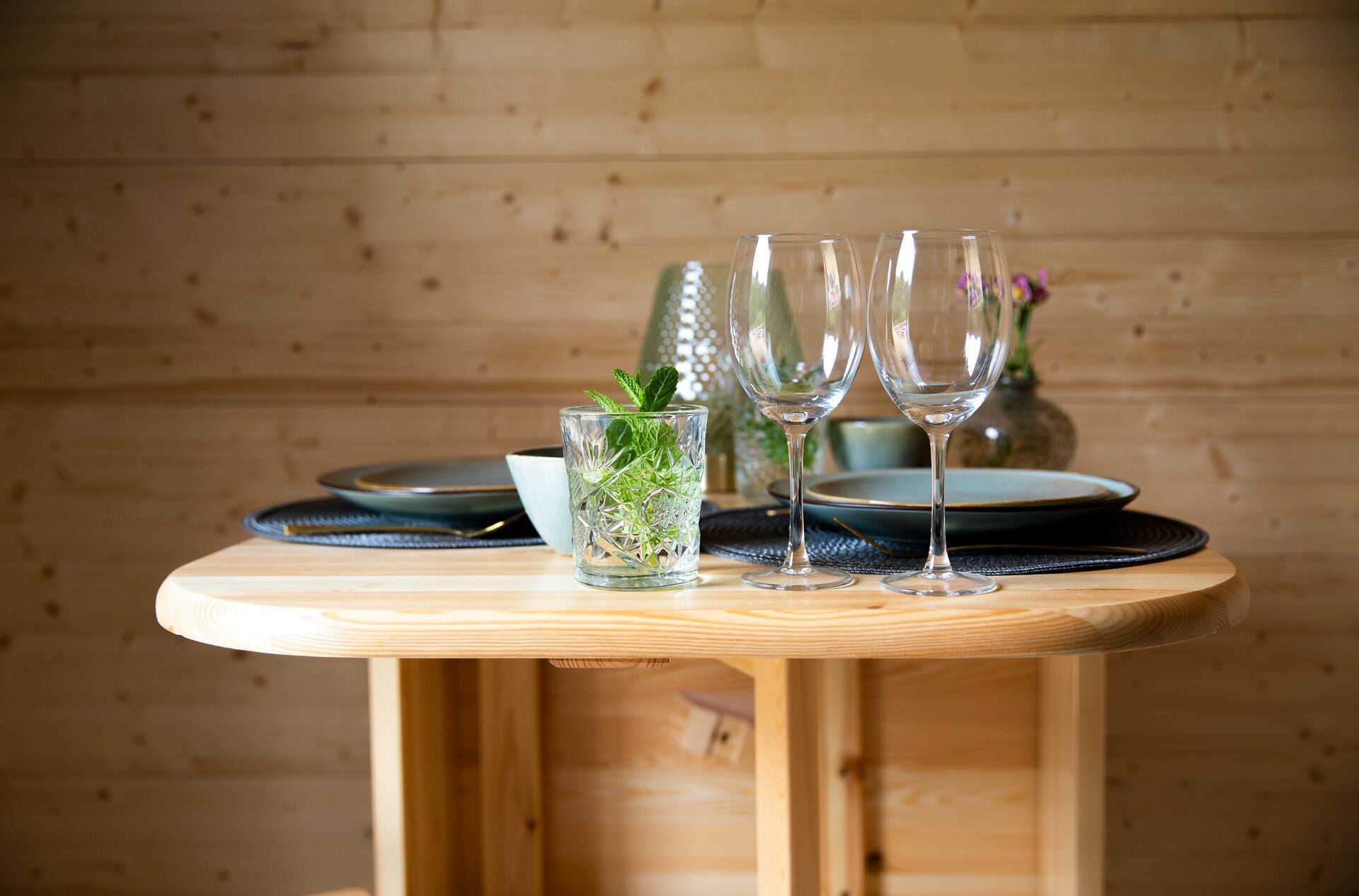 Una mesa de madera con tres vasos transparentes, un vaso con hojas de menta y una botella decorativa al fondo.