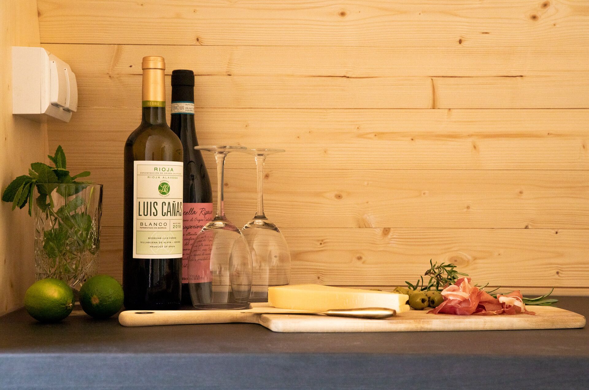 A bottle of wine, a glass, limes, cheese, olives, and prosciutto on a wooden cutting board.