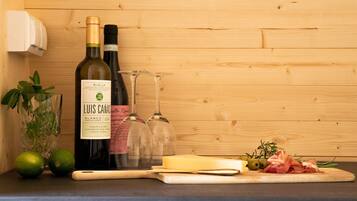 A bottle of wine, a glass, limes, cheese, olives, and prosciutto on a wooden cutting board.
