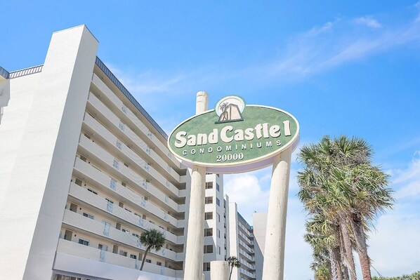 A sign for Sand Castle Condominiums with a clear view of a high-rise building in the background.