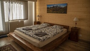 A double bed with patterned bedding, a wooden headboard, a nightstand with a lamp, a window with curtains, and a wall-mounted picture of a mountain landscape. - Great central location next to ski lifts and forest,spacious renovated apartment (Chamonix-Mont-Blanc)