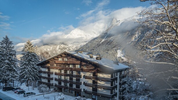 A multi-story hotel with a wooden facade, balconies, and a view of snow-covered mountains. - Great central location next to ski lifts and forest,spacious renovated apartment (Chamonix-Mont-Blanc)
