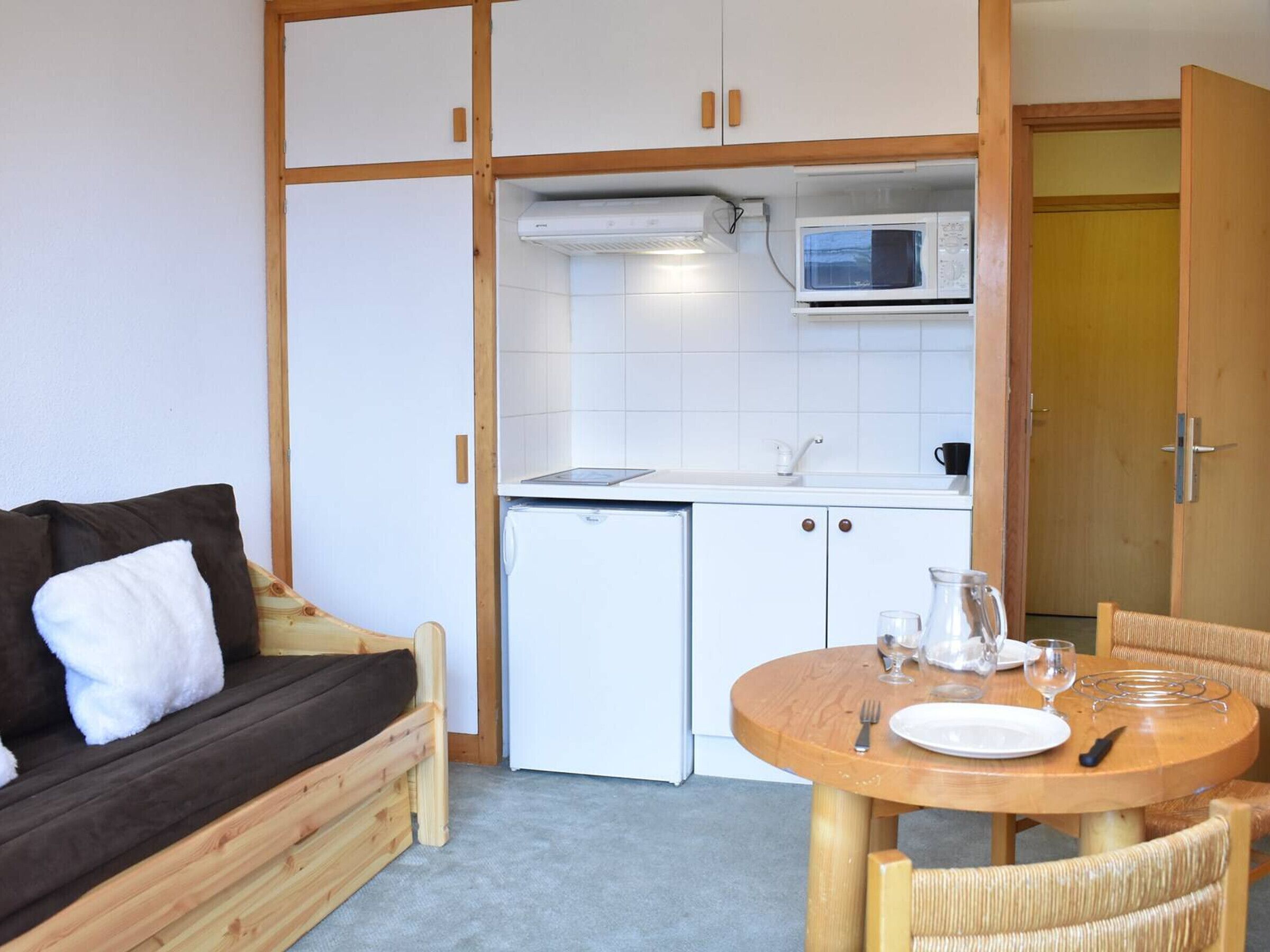 Una kitchenette compacta con microondas, un refrigerador pequeño y un lavamanos. Hay una mesa de madera con un plato, un vaso y un tenedor. Un sofá marrón con un cojín blanco está apoyado contra la pared.
