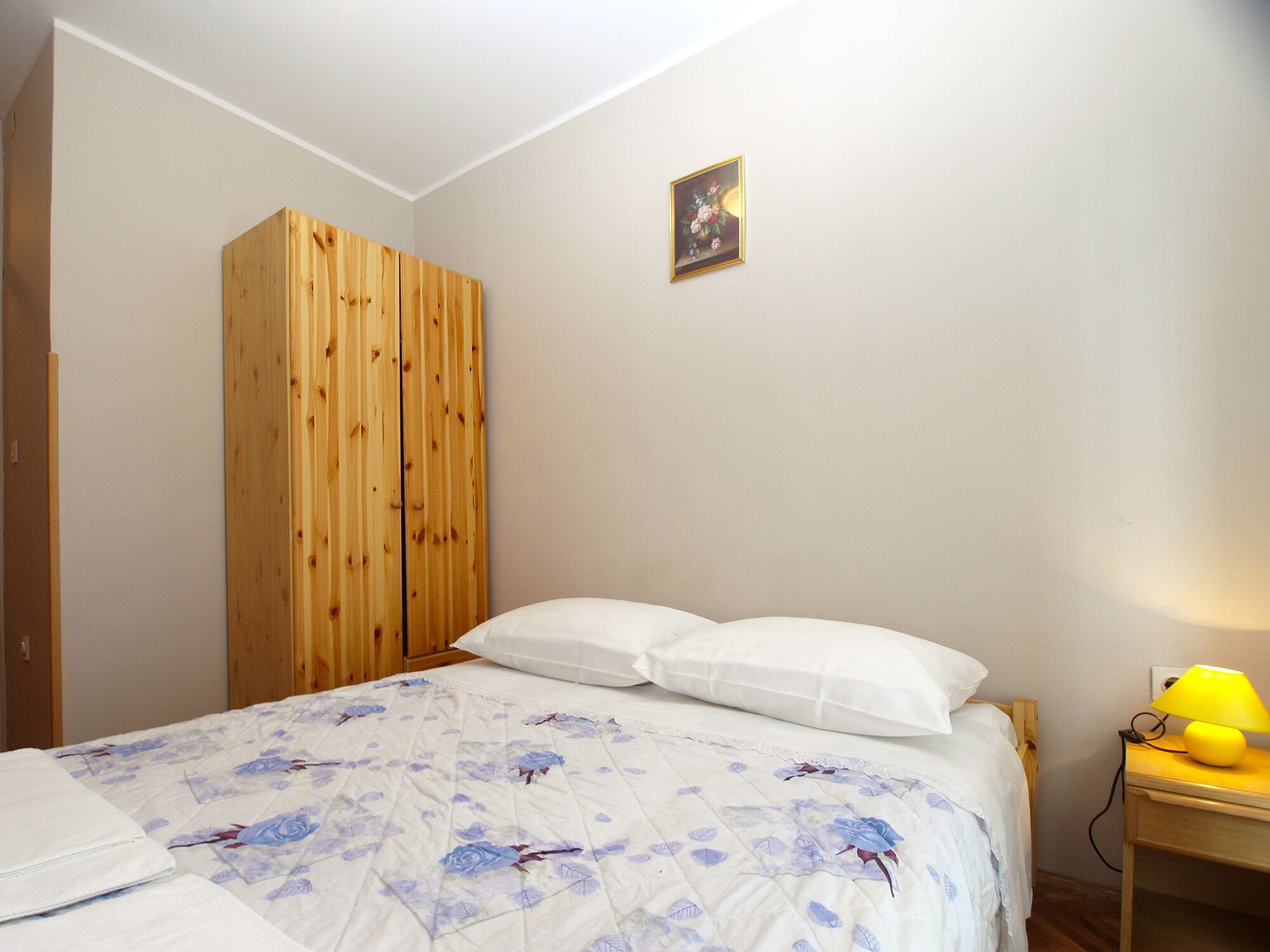 A bedroom with a bed, a wooden wardrobe, a nightstand with a lamp, and a small picture on the wall.