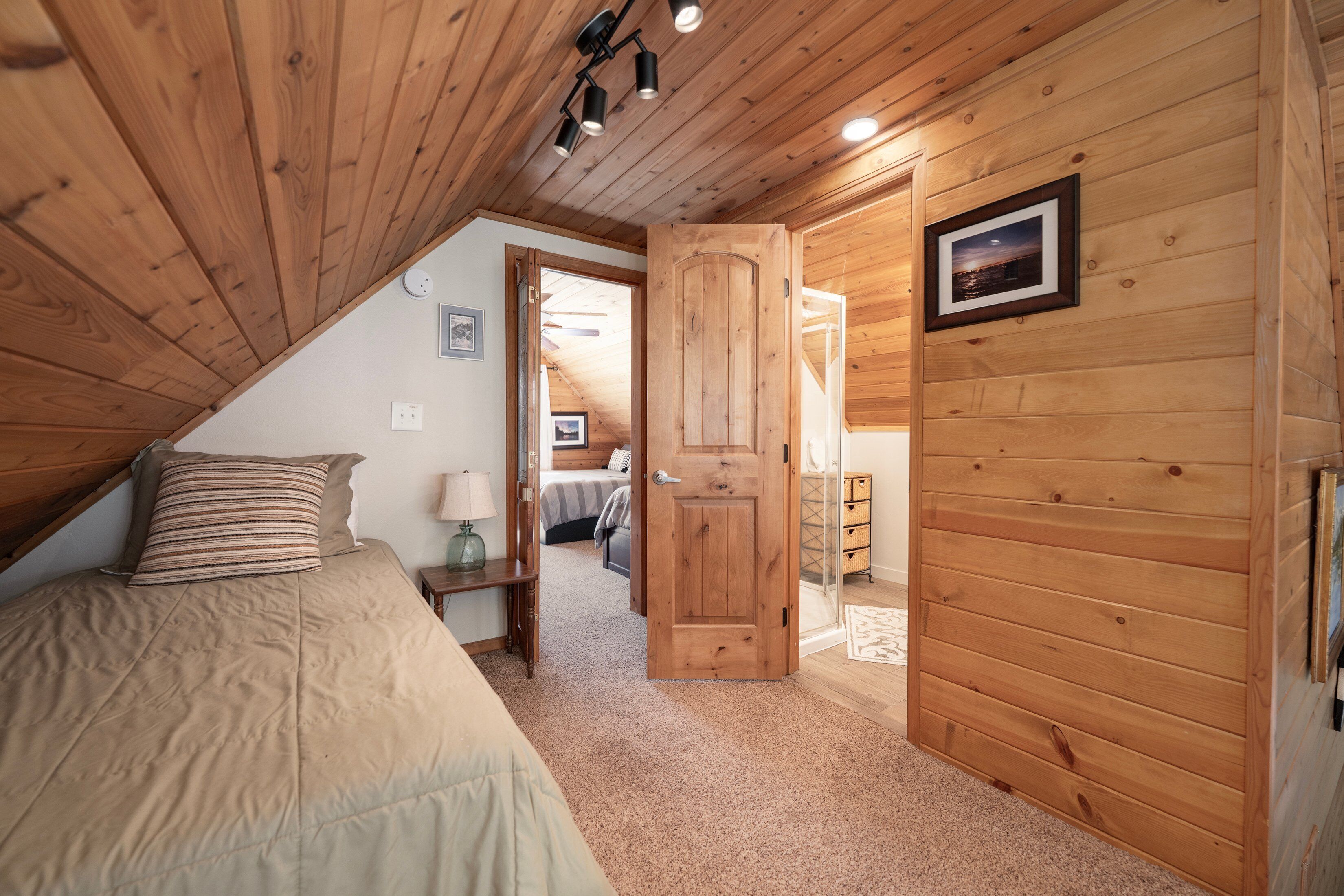 A bedroom with a wooden ceiling, a bed with a striped pillow, a nightstand with a lamp, and a door leading to another room.