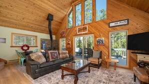 A cozy living room with a wooden ceiling, large windows, a fireplace, and a television.