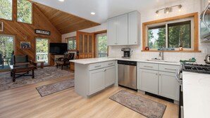 A modern kitchen with white cabinets, a stainless steel dishwasher, and a gas stove. There is a large window with a view of trees and a sign that reads "On Lake Time".
