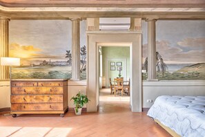 Una stanza con un letto, una cassettiera, una pianta in vaso e un affresco sulla parete.