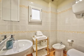 A bathroom with a sink, bidet, and toilet, a window, and a towel rack.