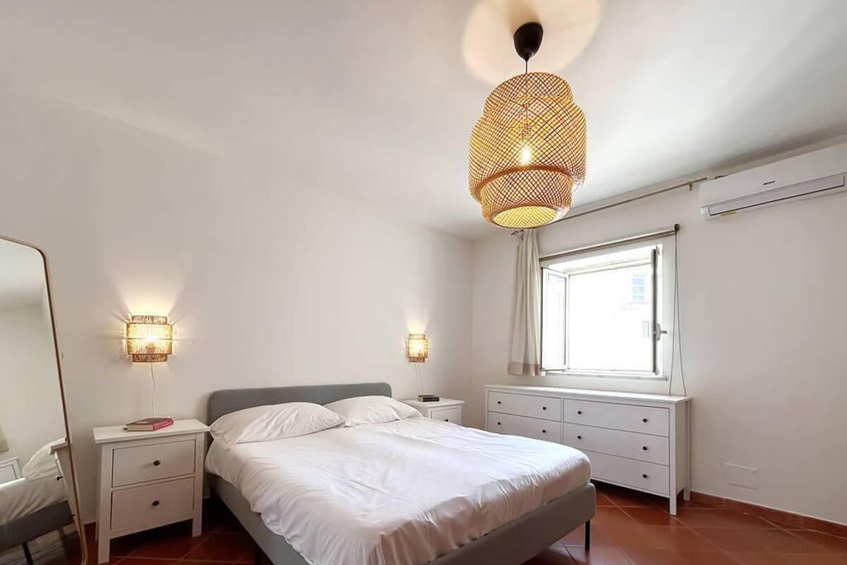 Un dormitorio con cama, mesitas de noche, un espejo, una ventana y una lámpara colgante.