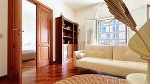 Una habitación con sofá, una estantería de madera, un tapiz de mimbre y una ventana con vistas a edificios.