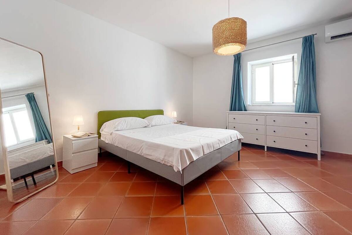 Un dormitorio con cama, mesitas de noche, cómoda, espejo y ventana con cortinas.