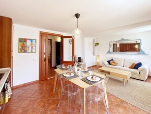 Un comedor con una mesa de madera dispuesta para cuatro personas, rodeada de sillas transparentes. Hay un sofá y una mesa auxiliar en la sala.