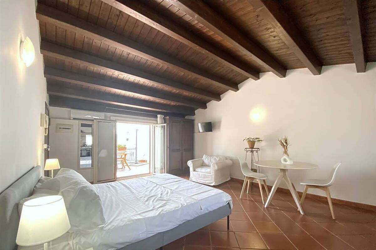 Un dormitorio con cama, lámpara de noche y una pequeña mesa con sillas. Hay una puerta que da a un balcón.