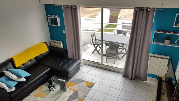 A living room with a black sofa, a glass coffee table, and a view of a patio with a table and chairs. - T1 bis Mezzanine, Très Lumineux, Proche Plage et Commodités (Le Château-d'Olonne)