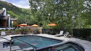 Une piscine avec un jacuzzi, entourée de meubles d’extérieur et de parasols, le tout adossé à un décor composé d’arbres et d’un bâtiment.