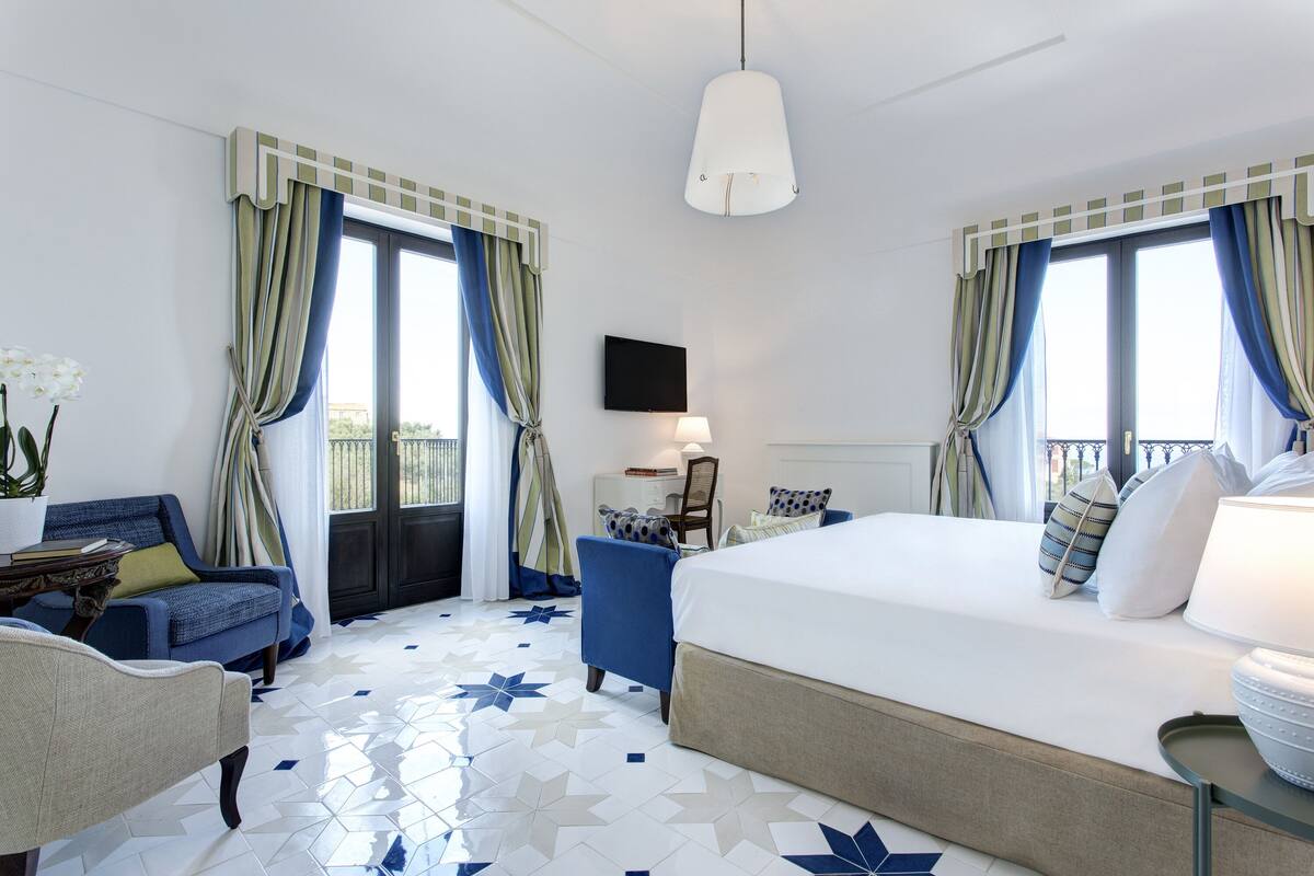A hotel room with a bed, a chair, a desk, a television, and a balcony with curtains.
