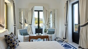 Quarto de hotel com uma cama, duas cadeiras, uma mesa e vista para o exterior através de uma grande janela.