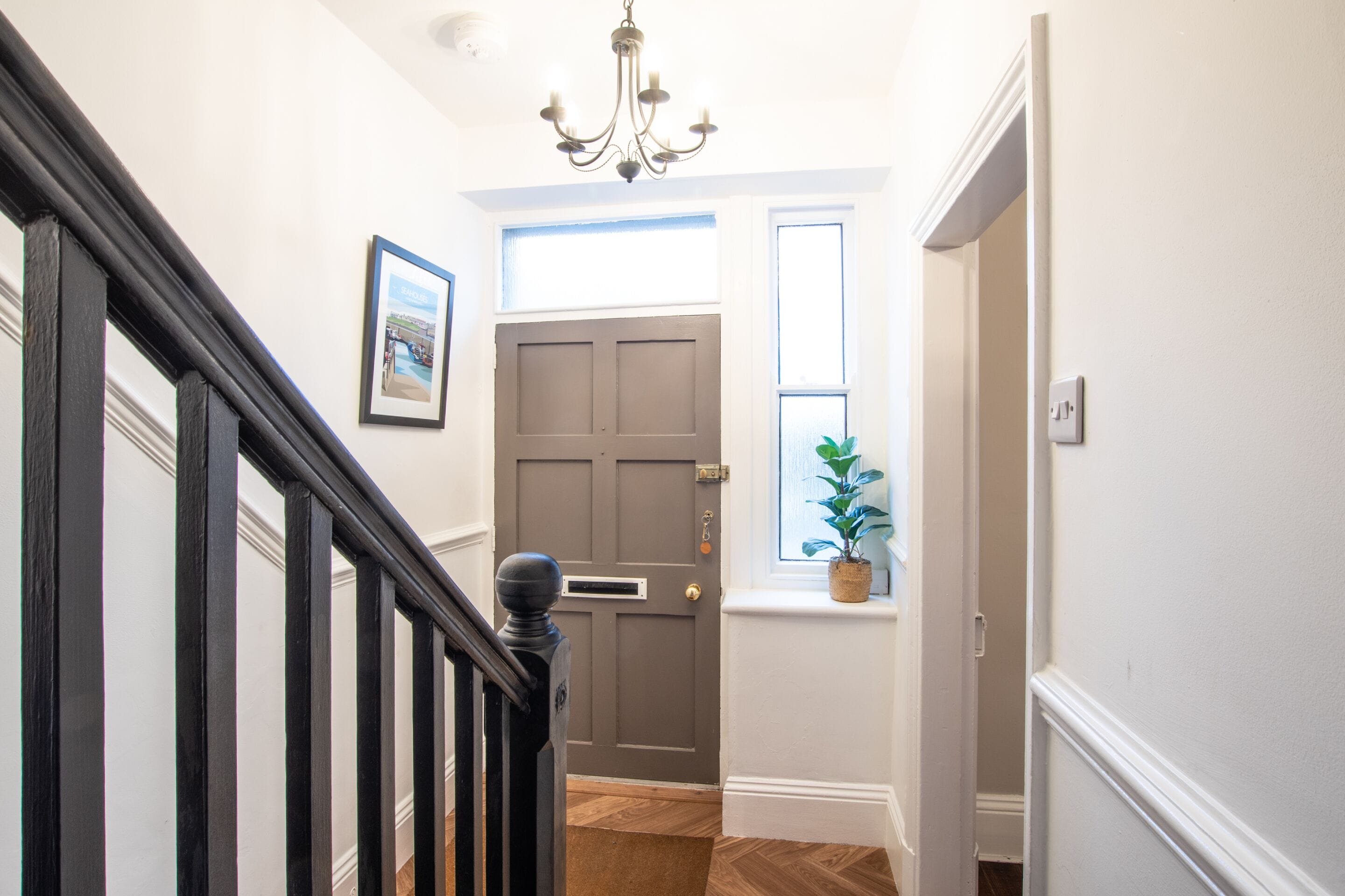 A hallway with a dark wooden staircase, a grey door, a window with a plant, and a chandelier.