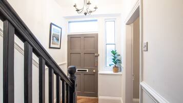 A hallway with a dark wooden staircase, a grey door, a window with a plant, and a chandelier.