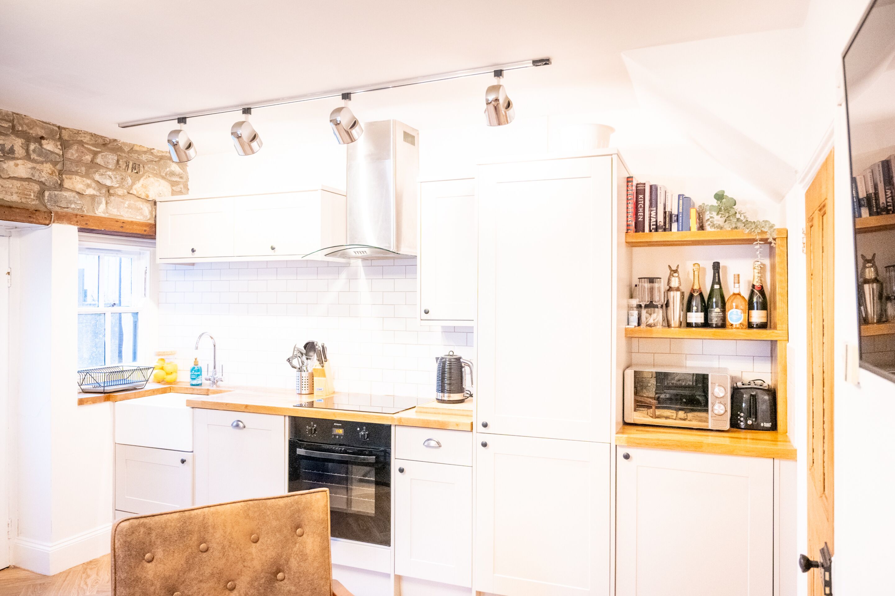 A modern kitchen with white cabinets, a wooden countertop, and a built-in oven. There is a bookshelf with books and bottles, and a stone wall in the background.