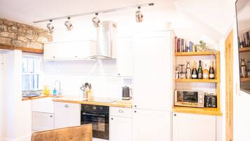 A modern kitchen with white cabinets, a wooden countertop, and a built-in oven. There is a bookshelf with books and bottles, and a stone wall in the background.